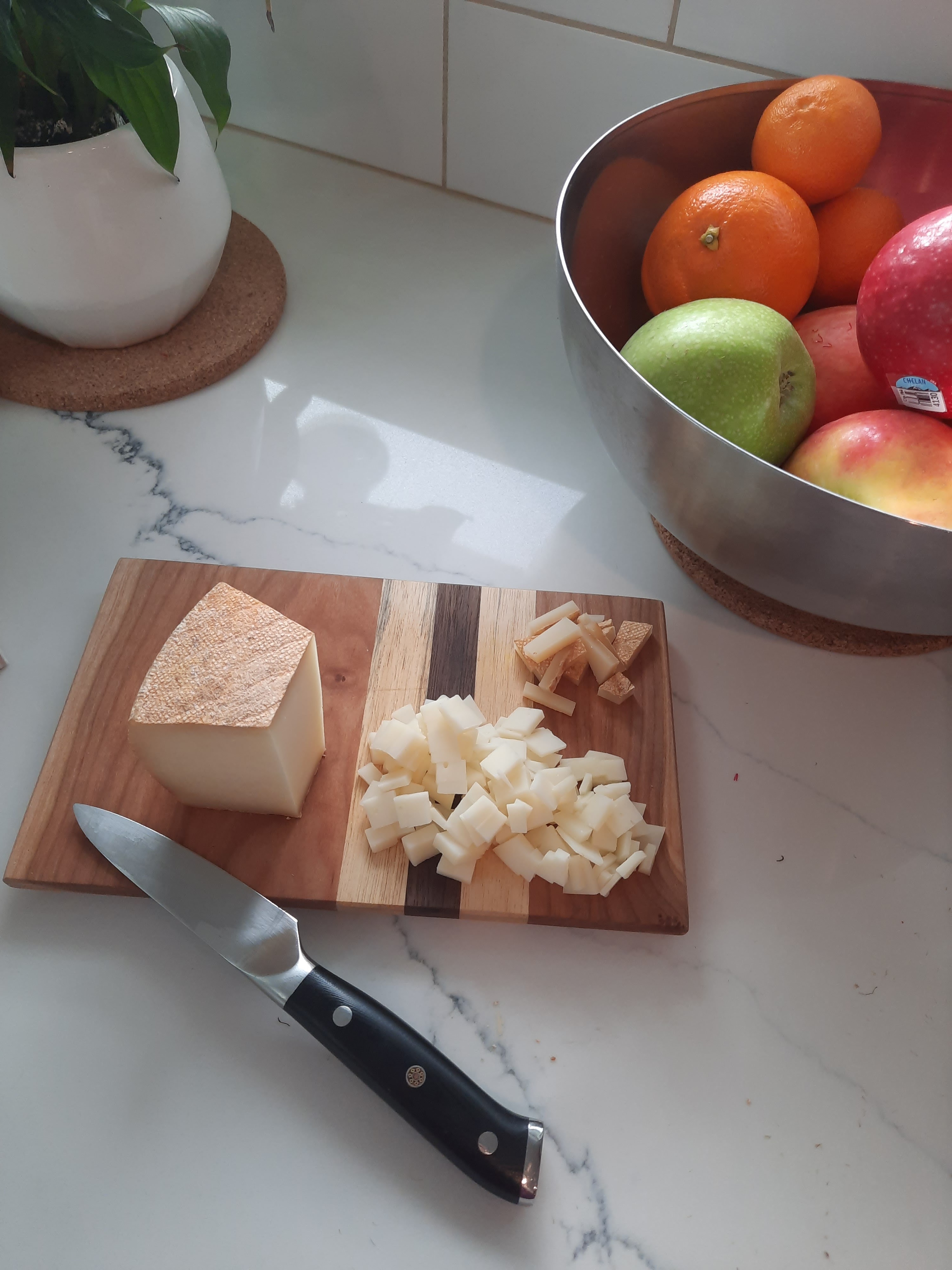 Ossau Iraty sheep milks ches chunk on a wooden cutting board. some pieces of cheese cut off into small cubes. small pairing knife lays on board as well. set against a marbled counter top with a plant and a bowl of fruit (apples and oranges) in the background